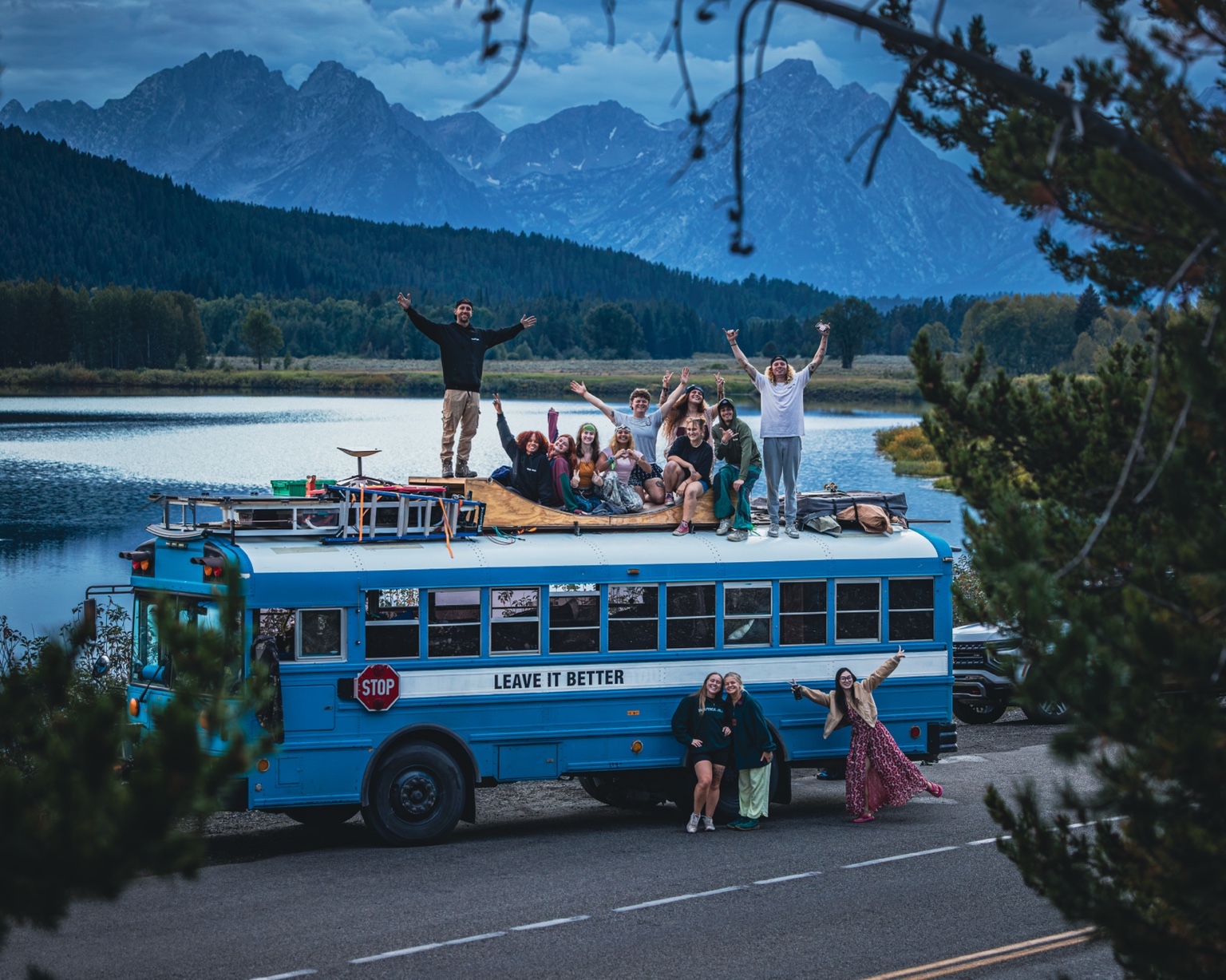 Theo and RoadTrippers on top of the blue school bus with mountains behind them